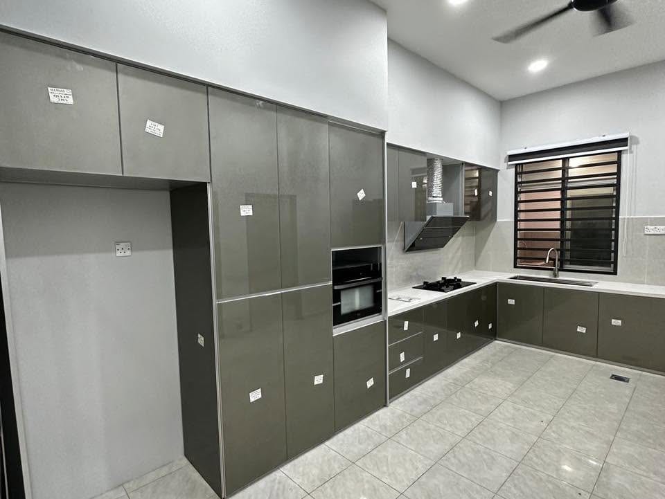 A newly installed L-shaped kitchen with olive-grey high-gloss doors, a tall unit housing a built-in oven and microwave, a chimney hood and white worktops, protective labels still on the doors.