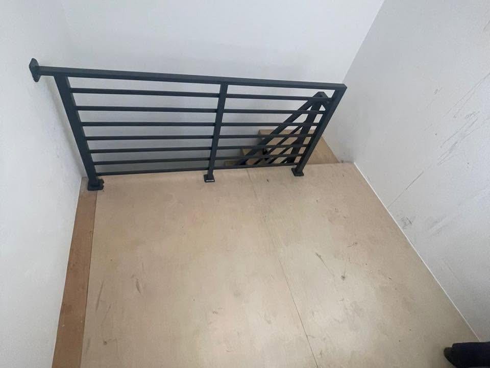 A dark grey horizontal-bar steel balustrade around a stair opening on a landing still covered with protective board, unpainted plaster walls.