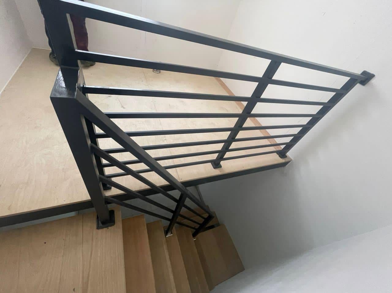Looking down a timber staircase onto a newly fitted dark grey horizontal-bar steel balustrade and landing rail, bare plaster walls, protective board on the landing.